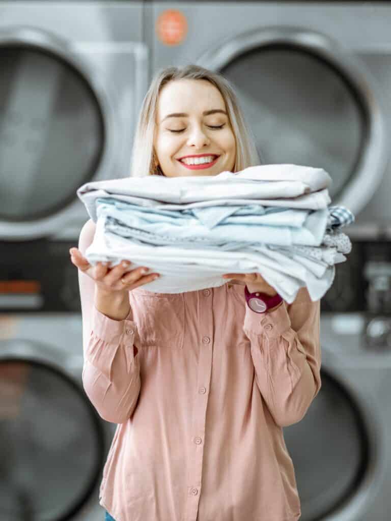 Junge Frau genießt sauber gebügelte Kleidung in der Selbstbedienungswäscherei mit Trocknern im Hintergrund.